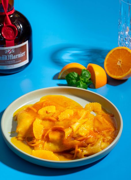 Eine Schale mit süßen Crêpes, umgeben von Orangenscheiben, Orangensauce und einer Flasche Grand Marnier Orangen-Likör.